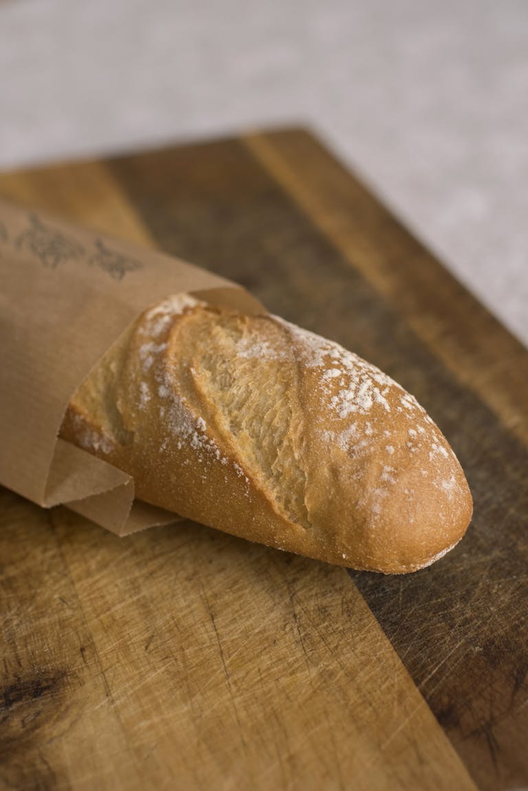 Freshly baked baguette wrapped in paper on a wooden breadboard, highlighting texture and warmth.